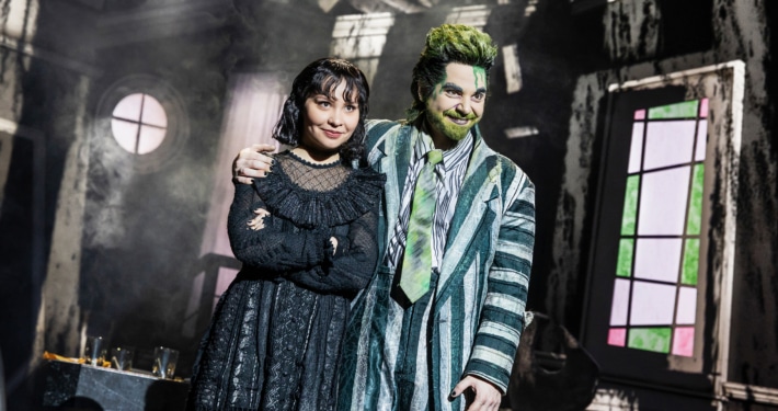 Two performers stand close together in a distressed interior set, one in a striped suit with green accents and one in a black gothic outfit, framed by a round window and angled walls.