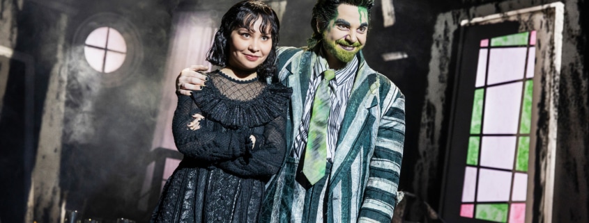Two performers stand close together in a distressed interior set, one in a striped suit with green accents and one in a black gothic outfit, framed by a round window and angled walls.