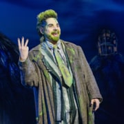 Performer in a striped suit with green accents stands onstage under blue lighting, raising three fingers while gesturing energetically; a dark, stylized set with a small house structure appears in the background.
