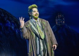 Performer in a striped suit with green accents stands onstage under blue lighting, raising three fingers while gesturing energetically; a dark, stylized set with a small house structure appears in the background.