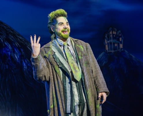 Performer in a striped suit with green accents stands onstage under blue lighting, raising three fingers while gesturing energetically; a dark, stylized set with a small house structure appears in the background.
