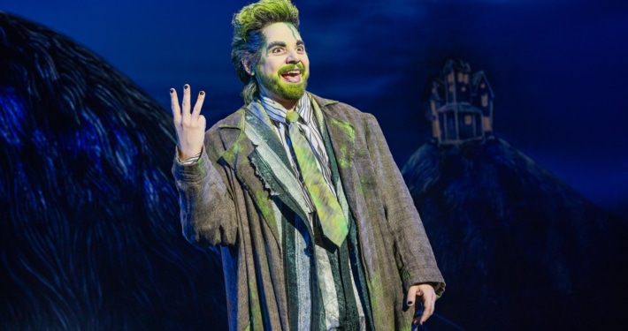 Performer in a striped suit with green accents stands onstage under blue lighting, raising three fingers while gesturing energetically; a dark, stylized set with a small house structure appears in the background.