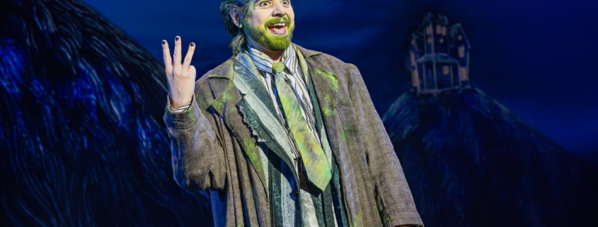 Performer in a striped suit with green accents stands onstage under blue lighting, raising three fingers while gesturing energetically; a dark, stylized set with a small house structure appears in the background.