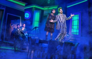 Four performers in a Beetlejuice stage scene: one in a striped suit stands on a table with arms open beside a performer in black, while two others react near a staircase; green and blue lighting with theatrical fog fills the room.