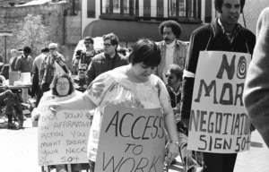 Historic black-and-white photograph of a disability rights demonstration. Several protesters, including wheelchair users, gather outdoors carrying signs with messages advocating for access to employment and the implementation of Section 504 protections.