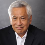 Professional headshot of a person with short gray hair wearing a dark suit jacket over a white collared shirt. The portrait is photographed against a neutral gray studio background with soft lighting and a professional business-style composition.