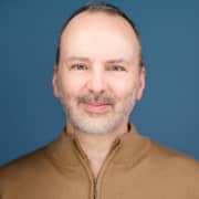 Close-cropped studio portrait of a person wearing a tan quarter-zip sweater against a blue background. The image focuses on the face and upper shoulders, with professional lighting and a simple backdrop.