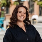 Outdoor portrait of a person with shoulder-length curly hair wearing a dark button-front blouse, standing on a pedestrian-friendly street with trees, benches, and softly blurred buildings in the background.