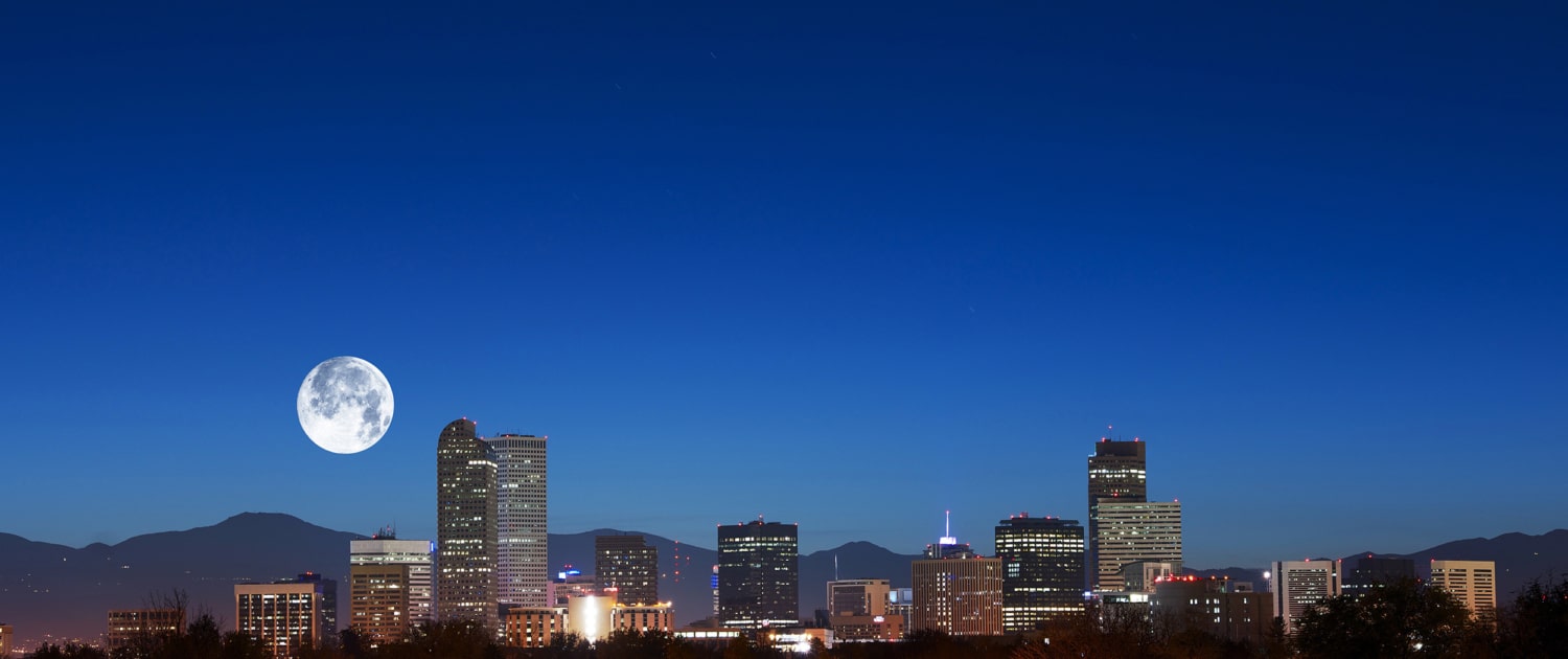 Nighttime view of the Denver skyline with illuminated high-rise buildings set against a deep blue sky. A large full moon appears above the mountains in the background, while trees, water, and glowing streetlights fill the foreground.