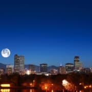 Nighttime view of the Denver skyline with illuminated high-rise buildings set against a deep blue sky. A large full moon appears above the mountains in the background, while trees, water, and glowing streetlights fill the foreground.