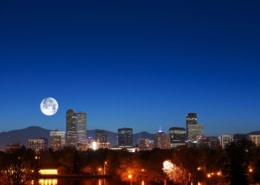 Nighttime view of the Denver skyline with illuminated high-rise buildings set against a deep blue sky. A large full moon appears above the mountains in the background, while trees, water, and glowing streetlights fill the foreground.