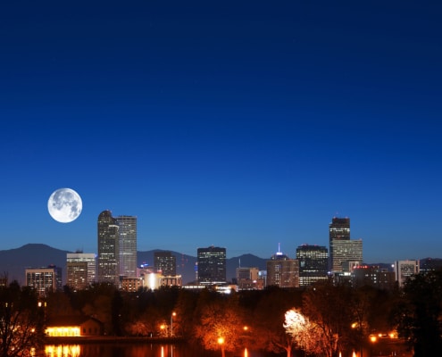 Nighttime view of the Denver skyline with illuminated high-rise buildings set against a deep blue sky. A large full moon appears above the mountains in the background, while trees, water, and glowing streetlights fill the foreground.