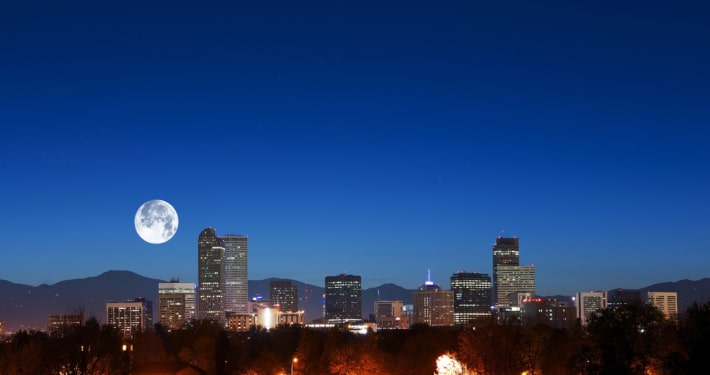 Nighttime view of the Denver skyline with illuminated high-rise buildings set against a deep blue sky. A large full moon appears above the mountains in the background, while trees, water, and glowing streetlights fill the foreground.