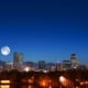 Nighttime view of the Denver skyline with illuminated high-rise buildings set against a deep blue sky. A large full moon appears above the mountains in the background, while trees, water, and glowing streetlights fill the foreground.