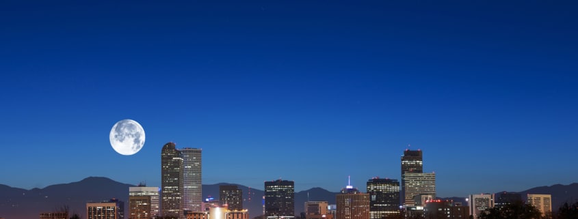 Nighttime view of the Denver skyline with illuminated high-rise buildings set against a deep blue sky. A large full moon appears above the mountains in the background, while trees, water, and glowing streetlights fill the foreground.