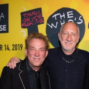 Two people pose together in front of a bright yellow promotional backdrop for The Who’s Tommy. One person wears a dark suit jacket with contrasting trim, and the other wears a dark collared shirt and necklace. Visible backdrop text includes “Tommy Can You Hear Me” and “The Who’s.”