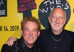 Two people pose together in front of a bright yellow promotional backdrop for The Who’s Tommy. One person wears a dark suit jacket with contrasting trim, and the other wears a dark collared shirt and necklace. Visible backdrop text includes “Tommy Can You Hear Me” and “The Who’s.”