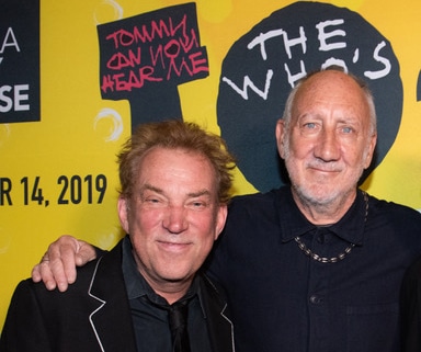 Two people pose together in front of a bright yellow promotional backdrop for The Who’s Tommy. One person wears a dark suit jacket with contrasting trim, and the other wears a dark collared shirt and necklace. Visible backdrop text includes “Tommy Can You Hear Me” and “The Who’s.”