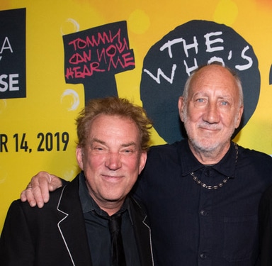 Two people pose together in front of a bright yellow promotional backdrop for The Who’s Tommy. One person wears a dark suit jacket with contrasting trim, and the other wears a dark collared shirt and necklace. Visible backdrop text includes “Tommy Can You Hear Me” and “The Who’s.”