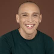 Professional headshot of a person with a shaved head wearing a dark green crew-neck shirt. The portrait is captured against a plain brown studio background with even lighting and a clean, minimalist composition.