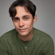 Professional headshot of a person with short gray hair wearing a dark suit jacket over a white collared shirt. The portrait is photographed against a neutral gray studio background with soft lighting and a professional business-style composition.