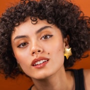 Professional headshot of a person with short curly hair wearing a black sleeveless top and star-shaped earrings. The portrait is photographed against a bright orange background with even studio lighting and a closely cropped composition.