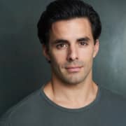 Professional headshot of a person with dark hair wearing a gray crew-neck shirt. The portrait is photographed against a dark blue-gray studio background with even lighting and a clean, centered composition.