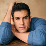 Professional portrait of a person wearing a textured blue long-sleeve shirt, posed with arms folded and head resting on one hand. The image is photographed against a warm beige background with soft, natural-looking lighting and a close-cropped composition.