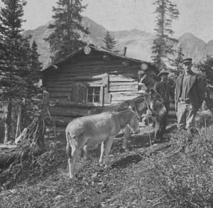 Historic black-and-white photograph showing several people standing beside a log cabin in a mountain setting. A donkey stands in the foreground, with tall pine trees and rugged peaks visible in the background.