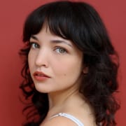 Professional headshot of a person with shoulder-length dark wavy hair and bangs, wearing a light-colored sleeveless top. The portrait is photographed against a solid red background with soft studio lighting and a close-cropped composition.