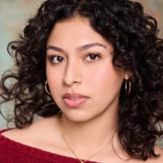 Professional headshot of a person with dark curly shoulder-length hair wearing a red knit top, hoop earrings, and a delicate necklace. The portrait is closely cropped and photographed against a softly blurred studio-style background with neutral tones.