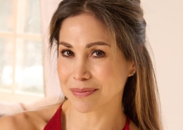 Professional headshot of a person wearing a red halter-style top, photographed indoors with soft natural light. The portrait features a neutral background with a window softly out of focus behind the subject.