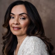 Professional headshot of a person with shoulder-length dark hair wearing a textured white top. The portrait is photographed against a warm brown studio background with soft lighting and a close-cropped composition.