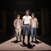 Production photo from The Outsiders showing three performers standing under a dramatic spotlight on a dark stage. Wearing casual, mid-century-inspired clothing including jeans, T-shirts, and a denim vest, the performers walk forward across a brightly lit section of the set while the surrounding stage remains in shadow.