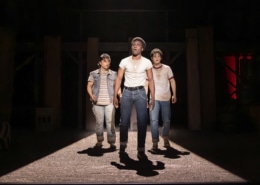Production photo from The Outsiders showing three performers standing under a dramatic spotlight on a dark stage. Wearing casual, mid-century-inspired clothing including jeans, T-shirts, and a denim vest, the performers walk forward across a brightly lit section of the set while the surrounding stage remains in shadow.