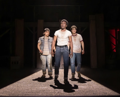 Production photo from The Outsiders showing three performers standing under a dramatic spotlight on a dark stage. Wearing casual, mid-century-inspired clothing including jeans, T-shirts, and a denim vest, the performers walk forward across a brightly lit section of the set while the surrounding stage remains in shadow.