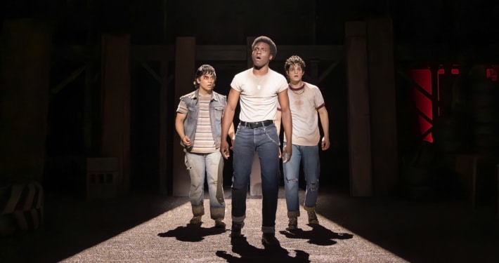 Production photo from The Outsiders showing three performers standing under a dramatic spotlight on a dark stage. Wearing casual, mid-century-inspired clothing including jeans, T-shirts, and a denim vest, the performers walk forward across a brightly lit section of the set while the surrounding stage remains in shadow.