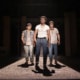 Production photo from The Outsiders showing three performers standing under a dramatic spotlight on a dark stage. Wearing casual, mid-century-inspired clothing including jeans, T-shirts, and a denim vest, the performers walk forward across a brightly lit section of the set while the surrounding stage remains in shadow.