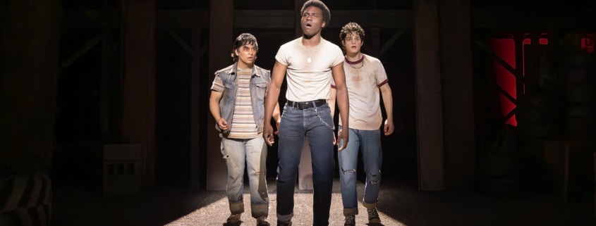 Production photo from The Outsiders showing three performers standing under a dramatic spotlight on a dark stage. Wearing casual, mid-century-inspired clothing including jeans, T-shirts, and a denim vest, the performers walk forward across a brightly lit section of the set while the surrounding stage remains in shadow.