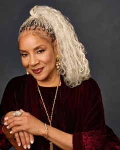 Studio portrait of a person with long light-colored textured hair, wearing a burgundy velvet top, pearl earrings, and gold jewelry, seated against a dark gray background.