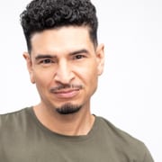 Professional headshot of a person with short curly hair wearing an olive-green crew-neck shirt. The portrait is photographed against a light gray studio background with a clean, minimalist composition.