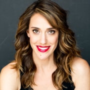 Professional headshot of a person with shoulder-length wavy hair wearing a black sleeveless top and bright red lipstick. The portrait is closely cropped against a dark studio background with even lighting.