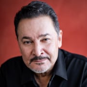 Professional headshot of a person with short dark hair and a neatly trimmed goatee, wearing a black collared shirt. The portrait is photographed against a warm red background with soft lighting and a close-cropped composition.