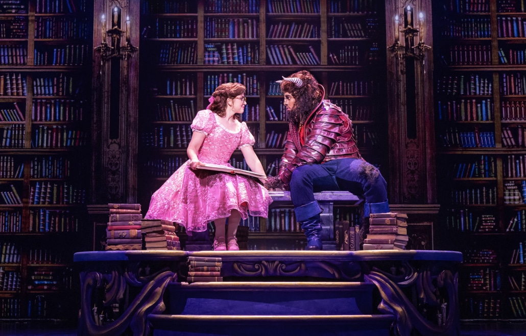 Belle and the Beast share a quiet moment in the castle library in Disney's Beauty and the Beast, surrounded by towering bookshelves.