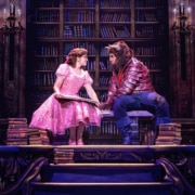 Belle and the Beast share a quiet moment in the castle library in Disney's Beauty and the Beast, surrounded by towering bookshelves.