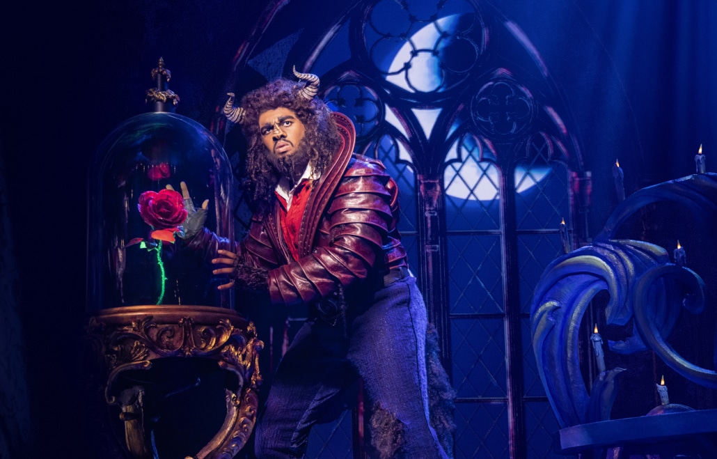 An image of an actor dressed as a beast with horns and facial hair standing in front of a glass-enclosed red rose