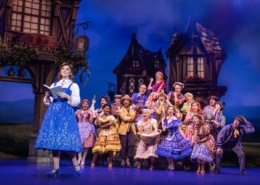 Belle reads a book as villagers gather behind her during the opening village scene in Disney's Beauty and the Beast.
