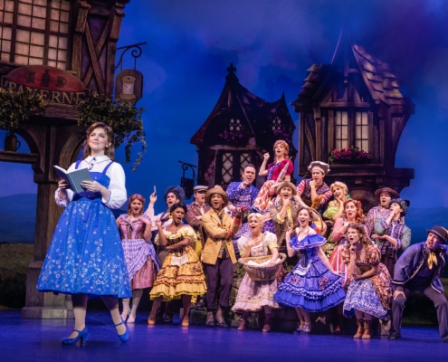 Belle reads a book as villagers gather behind her during the opening village scene in Disney's Beauty and the Beast.