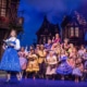 Belle reads a book as villagers gather behind her during the opening village scene in Disney's Beauty and the Beast.