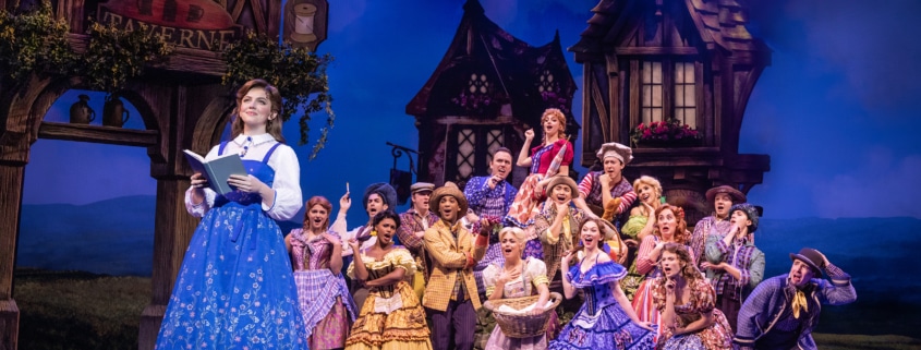 Belle reads a book as villagers gather behind her during the opening village scene in Disney's Beauty and the Beast.